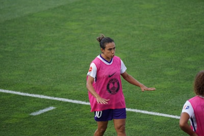 A soccer player is on a field wearing a pink training vest over a white and purple uniform. The player appears to be moving with focus and concentration. The grass is green and well-maintained.