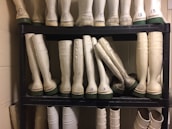 A neatly organized black shelf holds multiple pairs of tall rubber boots, primarily white with green soles. The boots are arranged in rows with some pairs labeled by numbers. The setting appears to be a utilitarian or industrial storage space with beige walls and some visible numbering on the boots.