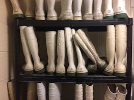 A neatly organized black shelf holds multiple pairs of tall rubber boots, primarily white with green soles. The boots are arranged in rows with some pairs labeled by numbers. The setting appears to be a utilitarian or industrial storage space with beige walls and some visible numbering on the boots.