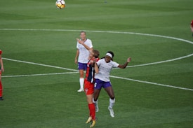 Two soccer players are competing for the ball in mid-air. One player is wearing a red and black uniform, while the other is in a white and purple kit. The background features a green soccer field.