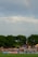 A soccer match is taking place with spectators sitting on a grassy hill behind a goalpost. A player in yellow is visible on the field. Various advertisements, including 'Nike Soccer', are displayed along the sidelines. Trees border the background, and a faint rainbow is visible in the cloudy sky.
