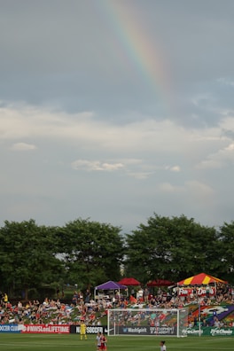 A soccer field bordered by green trees with a crowd of people gathered on a grassy area. Colorful tents are set up, with one displaying a sign that reads 'FAMILY DAY.' A rainbow is visible in the cloudy sky above.