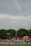 A soccer field bordered by green trees with a crowd of people gathered on a grassy area. Colorful tents are set up, with one displaying a sign that reads 'FAMILY DAY.' A rainbow is visible in the cloudy sky above.