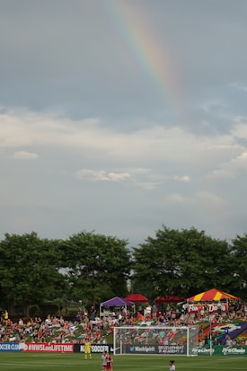 A soccer field bordered by green trees with a crowd of people gathered on a grassy area. Colorful tents are set up, with one displaying a sign that reads 'FAMILY DAY.' A rainbow is visible in the cloudy sky above.