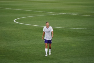A soccer player in a white and purple uniform standing on a grassy field, looking ahead with a neutral expression. The field markings are visible, indicating a professional sports setting.