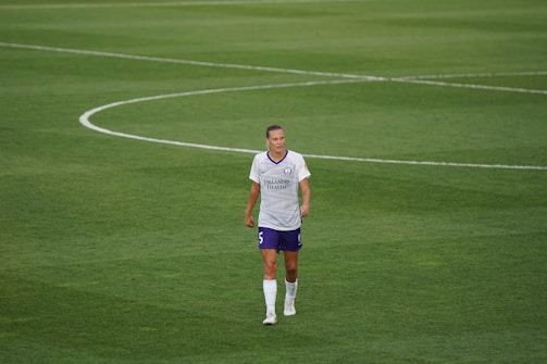 A soccer player in a white and purple uniform standing on a grassy field, looking ahead with a neutral expression. The field markings are visible, indicating a professional sports setting.