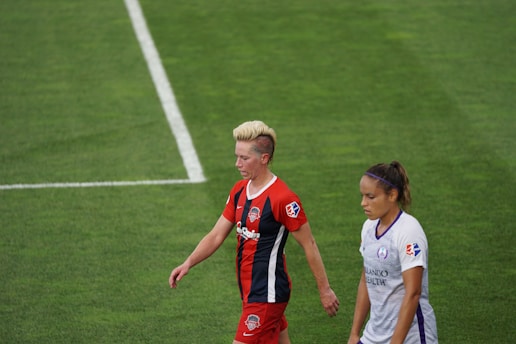 Two soccer players are walking on a grass field, one wearing a red and black striped jersey and the other in a white jersey. Both show focused expressions while an overhead perspective captures the in-game moment.