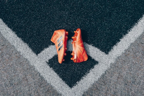 A pair of bright orange soccer cleats is placed on a textured black and gray surface, positioned symmetrically along intersecting white lines that form a V shape.