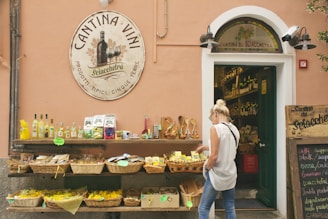A woman with blond hair is browsing a selection of goods displayed on shelves outside a store. The shop is adorned with a large round sign that reads 'Cantina Vini,' featuring images of wine bottles and glasses. Various products, including bottles, jars, and packaged goods, are organized in baskets and arranged neatly. The store entrance shows more items inside, indicating it is a specialized wine or local goods store.