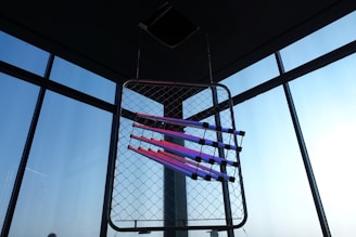 A geometric art installation is suspended in front of large windows. The installation consists of fluorescent light tubes in shades of pink and purple arranged diagonally across a wire mesh frame. The background features a clear blue sky visible through the windows.