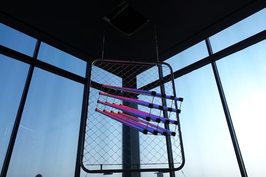 A geometric art installation is suspended in front of large windows. The installation consists of fluorescent light tubes in shades of pink and purple arranged diagonally across a wire mesh frame. The background features a clear blue sky visible through the windows.