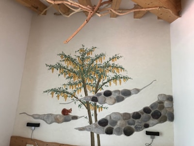 A mural painted on a wall depicts a tree with green leaves and yellow flowers. Large stones form an abstract pattern along the wall, and a small sculpture or painting of a brown squirrel is perched on one of the stone formations. The ceiling features exposed wooden beams and decorative branches.