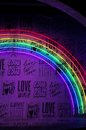 A wall covered in various affirming phrases related to love, featuring a prominent neon rainbow lighting installation. The words are bold and include messages such as 'love wins' and 'support love' in different typographies.