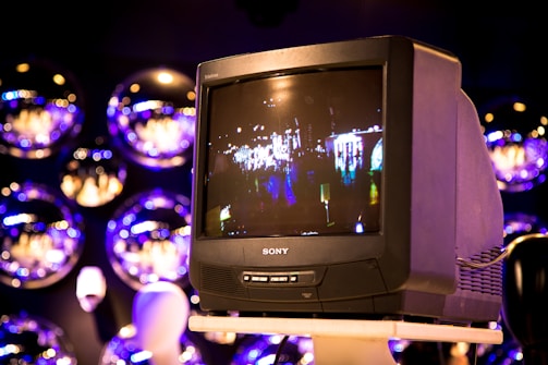 A vintage Sony Trinitron television is placed on a stand, displaying a blurred scene of lights and figures, possibly from a cityscape. The background features multiple reflective spheres, creating an artistic and modern ambiance.