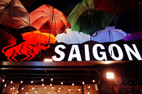 A collection of colorful umbrellas hangs from the ceiling, overlapping in hues of red, green, purple, and blue. Below them, there is a vibrant illuminated sign that reads 'SAIGON' accompanied by a stylized red bull logo. Decorative string lights are visible below the sign, adding a warm and inviting atmosphere.