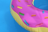 A colorful inflatable pool float designed to resemble a donut with pink icing and multicolored sprinkles floating on a clear blue water surface.
