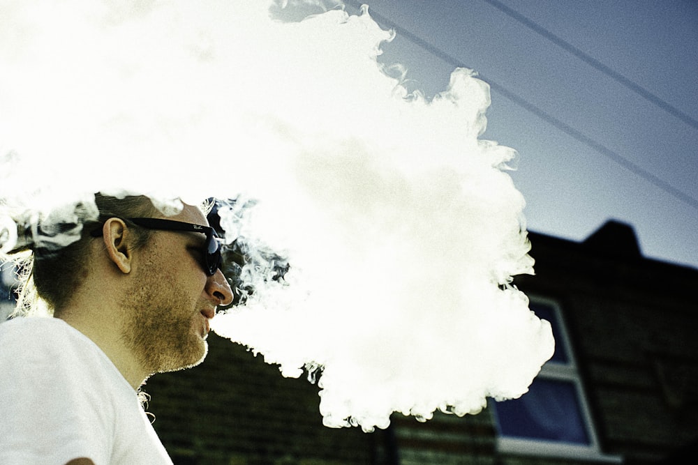 man blowing thick white smoke on air overlooking black building wall at daytime