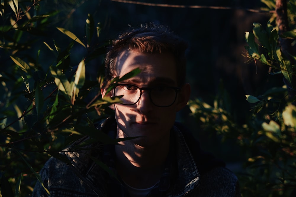 photography of man standing while wearing eyeglasses between near green leaf plants