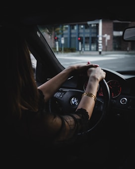 A person drives a BMW vehicle with a focus on the steering wheel and the driver's hands adorned with a bracelet. Outside, a city street with traffic lights is visible through the windshield.