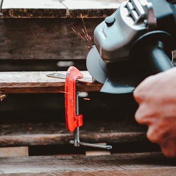 A close-up view of a hand-operated angle grinder cutting through a flat metal object secured with a red clamp. Sparks are visible as the grinder makes contact with the metal. The scene suggests a workshop or construction setting with wooden surfaces beneath.