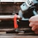 A close-up view of a hand-operated angle grinder cutting through a flat metal object secured with a red clamp. Sparks are visible as the grinder makes contact with the metal. The scene suggests a workshop or construction setting with wooden surfaces beneath.