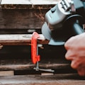 A close-up view of a hand-operated angle grinder cutting through a flat metal object secured with a red clamp. Sparks are visible as the grinder makes contact with the metal. The scene suggests a workshop or construction setting with wooden surfaces beneath.
