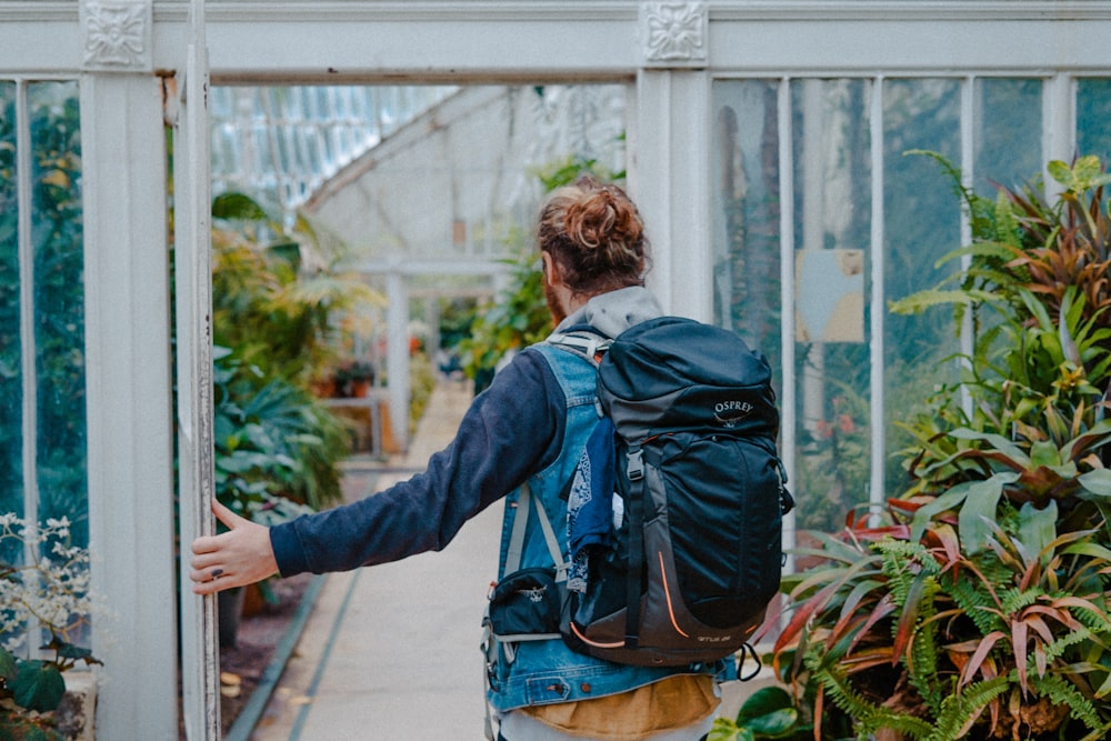 man in blue denim jacket and black hiking backpack stands on door near green plants
