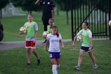 A group of individuals on a grassy field, with some wearing sports attire including a white and purple jersey. One person is holding a soccer ball, and others are wearing bright green vests with shorts. A man in the background has a whistle, suggesting a coaching role.