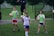 A group of individuals on a grassy field, with some wearing sports attire including a white and purple jersey. One person is holding a soccer ball, and others are wearing bright green vests with shorts. A man in the background has a whistle, suggesting a coaching role.