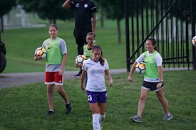 A group of individuals on a grassy field, with some wearing sports attire including a white and purple jersey. One person is holding a soccer ball, and others are wearing bright green vests with shorts. A man in the background has a whistle, suggesting a coaching role.
