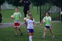 A group of individuals on a grassy field, with some wearing sports attire including a white and purple jersey. One person is holding a soccer ball, and others are wearing bright green vests with shorts. A man in the background has a whistle, suggesting a coaching role.