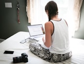 woman in white tank top sitting on bed in front of laptop computer