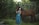 A woman in a flowing, colorful dress stands beside a brown horse in a lush, green forest. They appear to be sharing a peaceful moment, with the woman gently holding the horse's bridle as she looks up at it. Dense foliage and fallen branches surround them, creating a serene, natural setting.