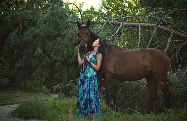 A woman in a flowing, colorful dress stands beside a brown horse in a lush, green forest. They appear to be sharing a peaceful moment, with the woman gently holding the horse's bridle as she looks up at it. Dense foliage and fallen branches surround them, creating a serene, natural setting.