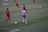 A women's soccer match is underway on a grass field. Players in red and black striped uniforms and others in white and blue uniforms are actively engaged in the game, with two players focused on controlling the ball near them.