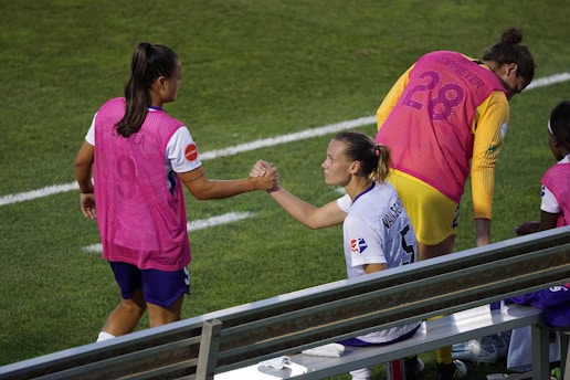 Two athletes wearing sports uniforms and pink vests engage in a fist bump on the sidelines of a sports field. The grassy field and metal bench seating are visible. One athlete is seated while the other stands next to them. Their expressions convey camaraderie and teamwork as they interact.