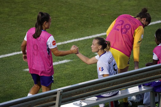 Two athletes wearing sports uniforms and pink vests engage in a fist bump on the sidelines of a sports field. The grassy field and metal bench seating are visible. One athlete is seated while the other stands next to them. Their expressions convey camaraderie and teamwork as they interact.