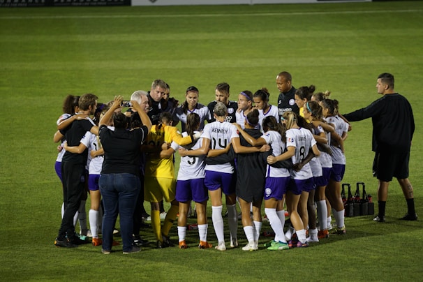 A group of soccer players and staff huddle together on a field, forming a tight circle. They are dressed in uniforms with some individuals in white and purple jerseys, while others wear black or yellow. The atmosphere seems focused and possibly motivational.