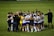A group of soccer players and staff huddle together on a field, forming a tight circle. They are dressed in uniforms with some individuals in white and purple jerseys, while others wear black or yellow. The atmosphere seems focused and possibly motivational.