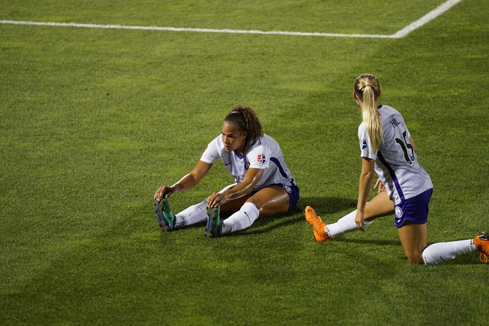 two women soccer players stretching legs on lawn