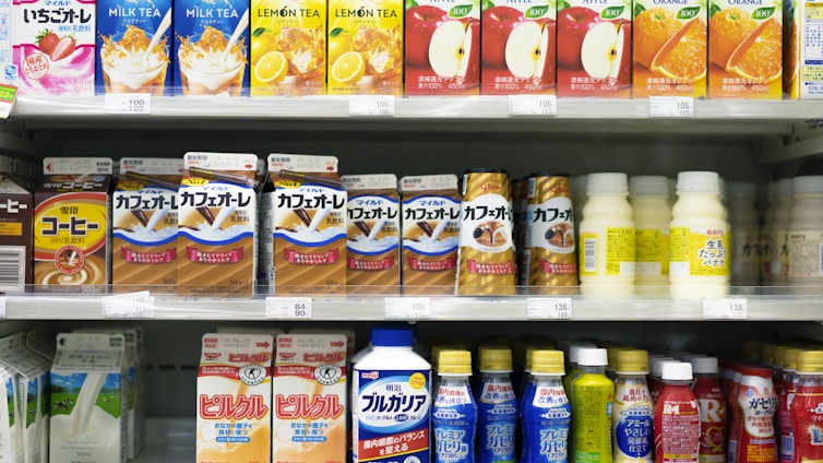 A supermarket shelf displays a variety of packaged drinks, including milk tea, lemon tea, apple juice, orange juice, coffee drinks, and other flavored beverages. The packaging features vibrant colors and images of ingredients such as fruits and coffee beans. Each product is neatly arranged in rows with price tags visible.