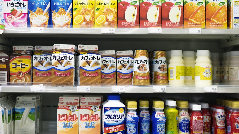 A supermarket shelf displays a variety of packaged drinks, including milk tea, lemon tea, apple juice, orange juice, coffee drinks, and other flavored beverages. The packaging features vibrant colors and images of ingredients such as fruits and coffee beans. Each product is neatly arranged in rows with price tags visible.