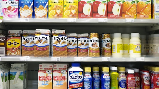 A supermarket shelf displays a variety of packaged drinks, including milk tea, lemon tea, apple juice, orange juice, coffee drinks, and other flavored beverages. The packaging features vibrant colors and images of ingredients such as fruits and coffee beans. Each product is neatly arranged in rows with price tags visible.