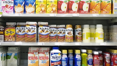A supermarket shelf displays a variety of packaged drinks, including milk tea, lemon tea, apple juice, orange juice, coffee drinks, and other flavored beverages. The packaging features vibrant colors and images of ingredients such as fruits and coffee beans. Each product is neatly arranged in rows with price tags visible.