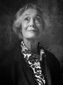 An elderly person with short, light-colored hair is looking slightly upwards with a thoughtful and serene expression. The person is wearing a patterned shirt with a floral design under a dark jacket. The background is a textured, dark wall.