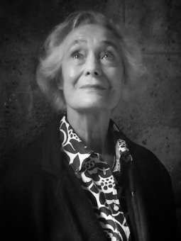 An elderly person with short, light-colored hair is looking slightly upwards with a thoughtful and serene expression. The person is wearing a patterned shirt with a floral design under a dark jacket. The background is a textured, dark wall.
