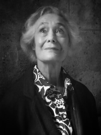 An elderly person with short, light-colored hair is looking slightly upwards with a thoughtful and serene expression. The person is wearing a patterned shirt with a floral design under a dark jacket. The background is a textured, dark wall.