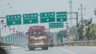 A highway scene with several green overhead road signs in both Chinese and English, indicating directions for different expressways and a bridge. A large truck is traveling on the road beneath the signs. Red lanterns hang from lampposts along the road, and trees line the highway. In the background, there are telecommunications towers.