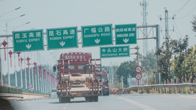 A highway scene with several green overhead road signs in both Chinese and English, indicating directions for different expressways and a bridge. A large truck is traveling on the road beneath the signs. Red lanterns hang from lampposts along the road, and trees line the highway. In the background, there are telecommunications towers.