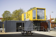 A structure made from shipping containers is set up outdoors. The lower container is dark gray and functions as the main space, featuring a door and large windows. On top sits a yellow container with large, clear windows, likely serving as an additional room or office space. A picnic table and a bicycle are located in front of the structure. Tall trees and a brick building are visible in the background under a clear sky.
